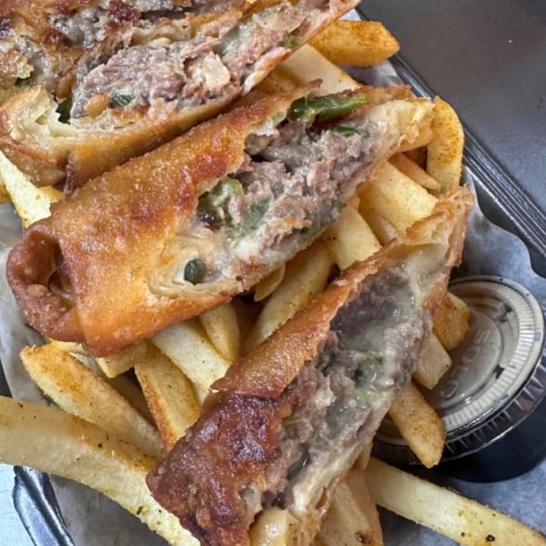 Cheesesteak egg rolls, cut open to show the filling, served with golden french fries and a dipping sauce.