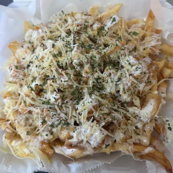 Golden french fries topped with white sauce, shredded cheese, and green herbs in a white container.