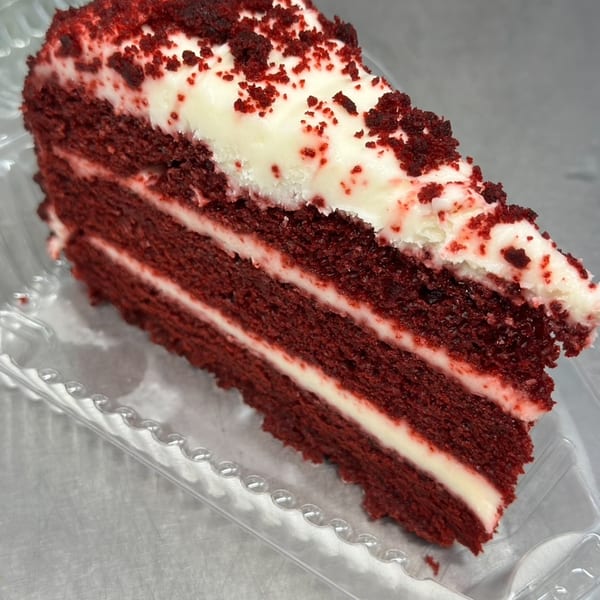 A slice of red velvet cake with cream cheese frosting and red crumbs in a plastic container.