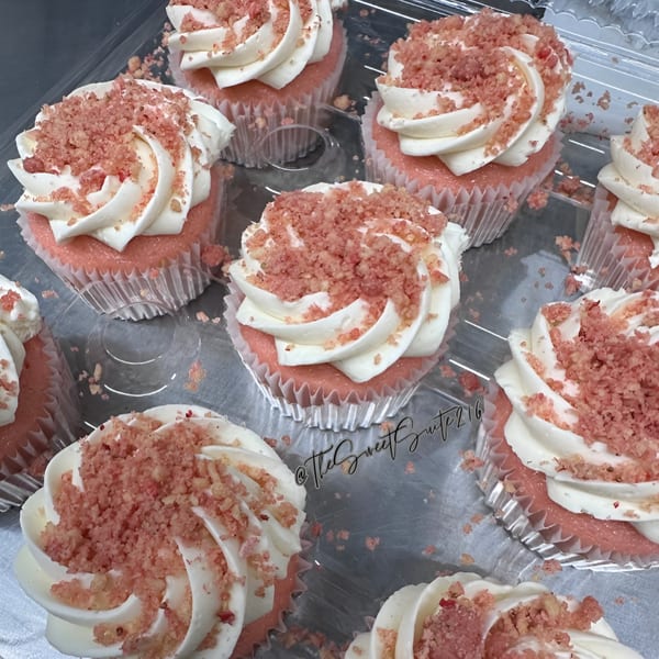 Pink cupcakes with white frosting and pink crumble topping in a clear plastic container.